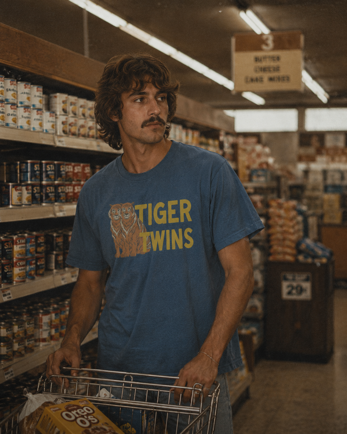 Tiger Twins tee in denim blue — mustard type with orange tiger duo, worn in a 1974 grocery aisle.